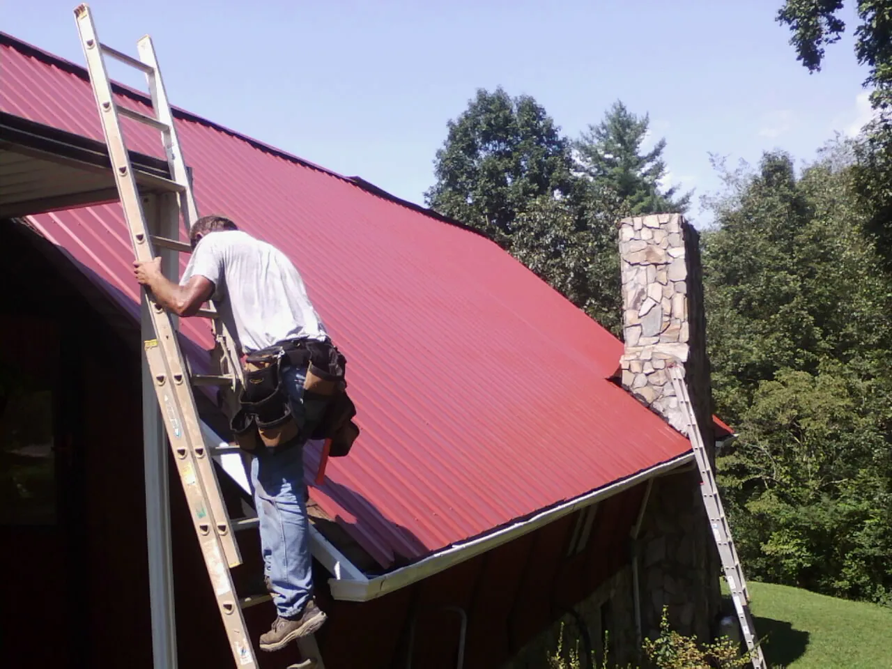 Roofer on a ladder beside a red metal roof and stone chimney — Soffit & Fascia Repair by Roofing Brothers in Peak